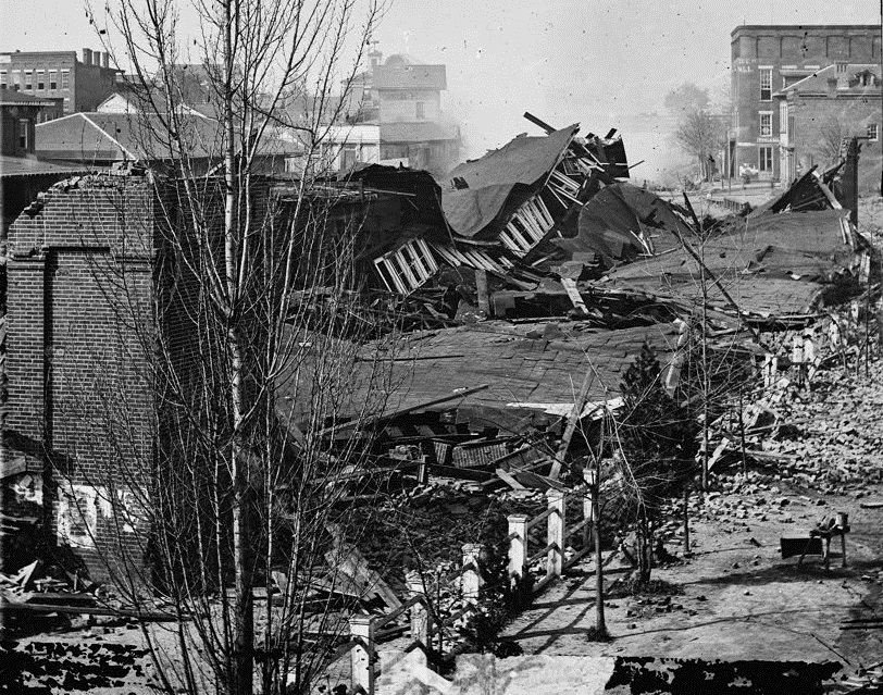 Black and white photograph of a collapsed railway depot, its roof caved in among brick rubble and bare trees.