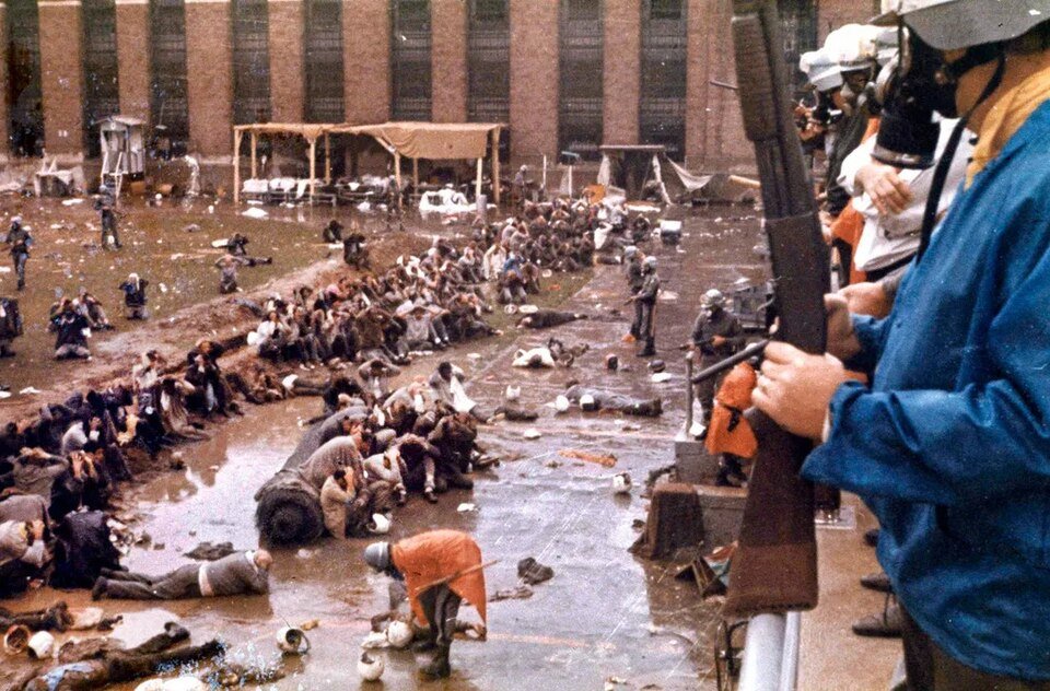 Colour photograph of a prison yard where large numbers of men sit and lie on wet ground watched by armed officers.