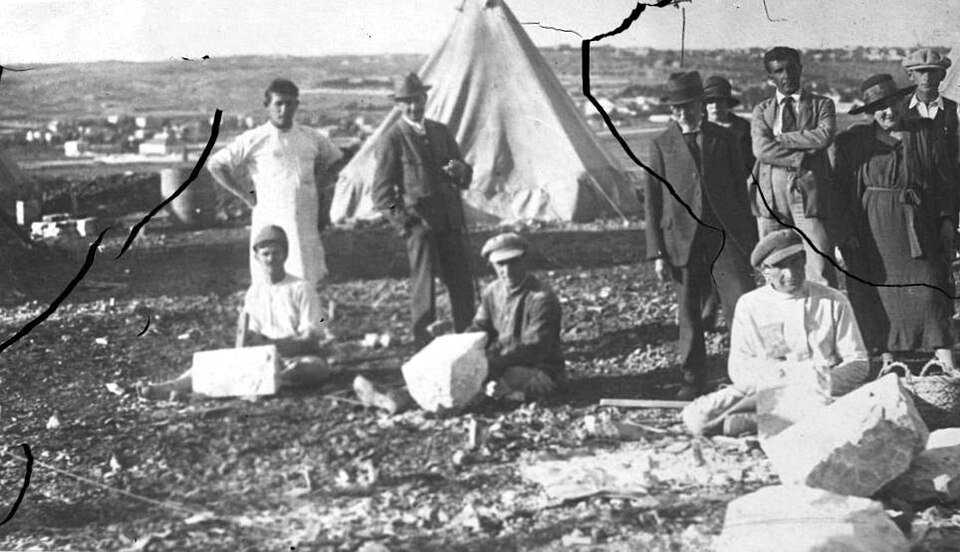 Damaged black and white photograph of men in work clothes and suits gathered on stony ground by a tent.