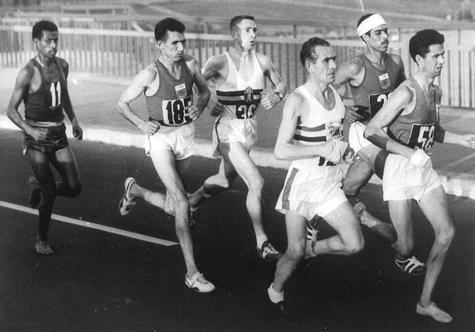A pack of six Olympic marathon runners on a road at night, the leader on the left running with bare feet while the others wear shoes.