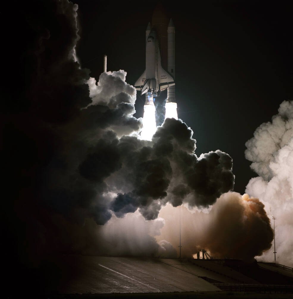 Photograph of a space shuttle lighting up the night sky with brilliant engine flames and huge billowing smoke clouds.