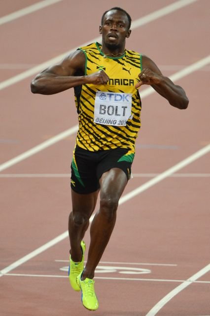Color photograph of a sprinter in a yellow and green Jamaica singlet crossing the finish line with arms raised.