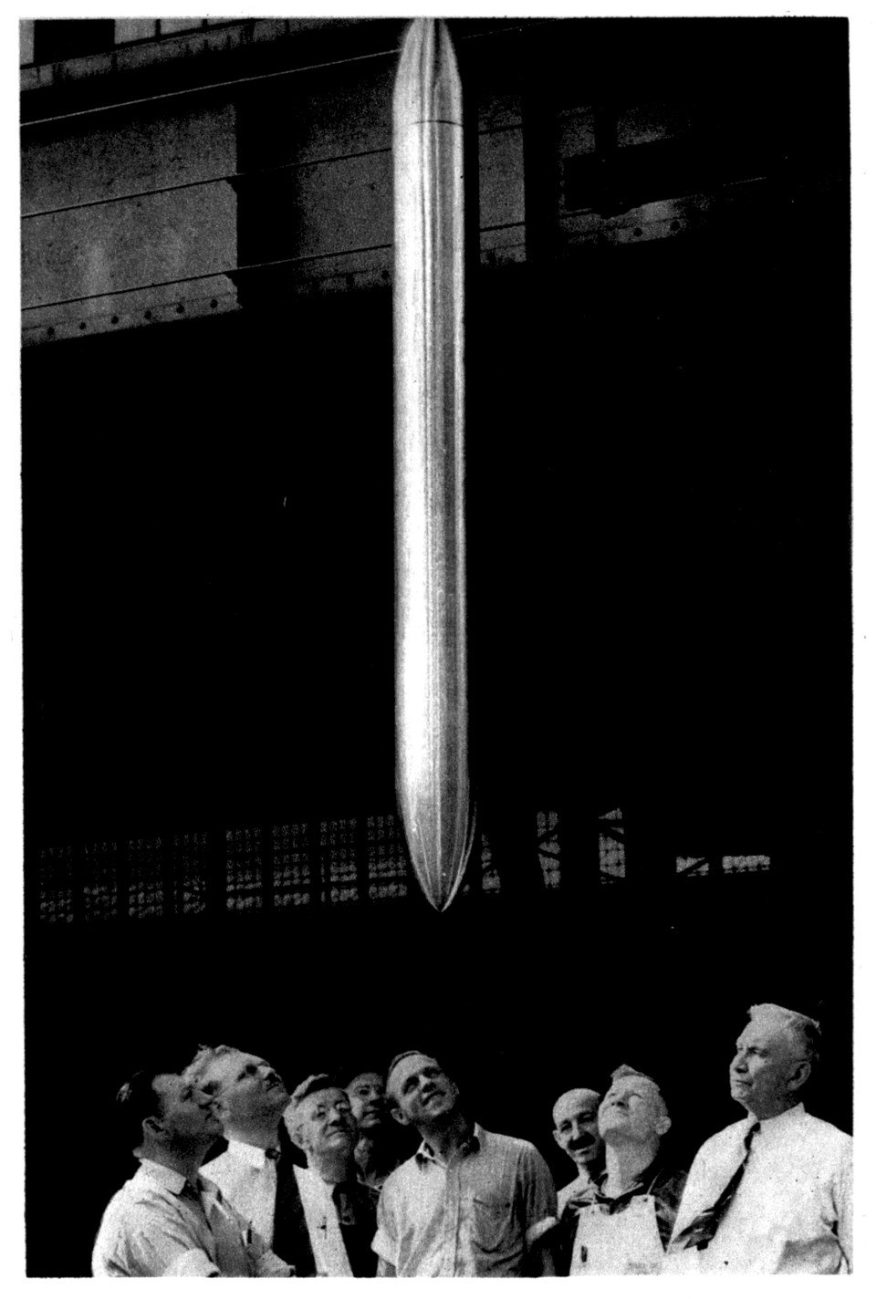 A long polished metal cylinder shaped like a torpedo hanging point-down, with a group of workmen in shirtsleeves gazing up at it.