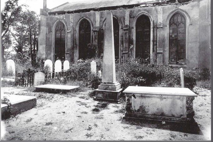 Black and white photograph of a damaged stone church with cracked walls, seen across an overgrown graveyard.