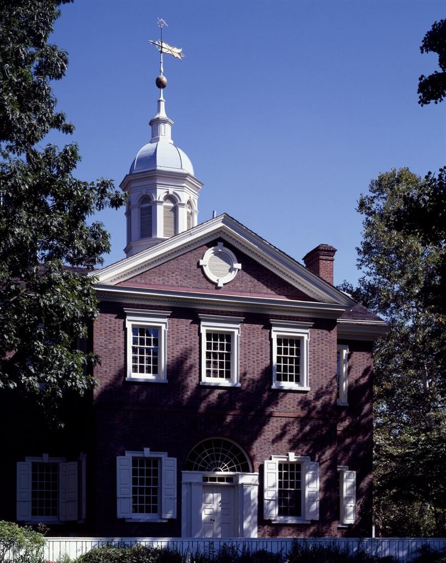 Colour photograph of a red brick Georgian hall with a white cupola and weathervane, framed by trees.