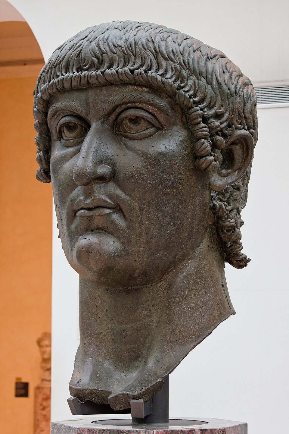 A colossal bronze head of a clean-shaven man with heavy-lidded eyes and a fringe of curls, displayed on a plinth in a gallery.