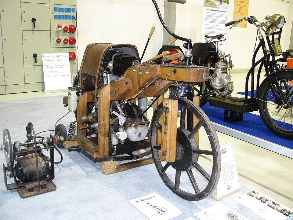 Photograph of an old wooden-framed motorized vehicle with a large rear wheel and visible engine, on display indoors.
