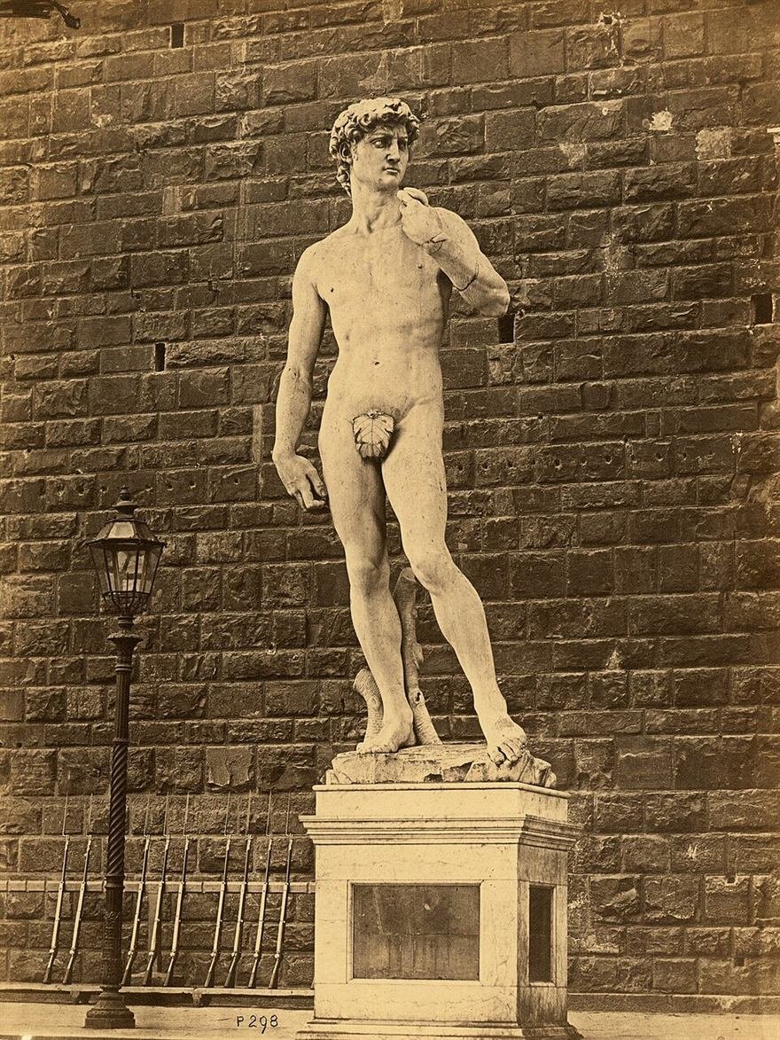 Sepia photograph of a colossal marble nude male statue on a plinth before a rough stone wall.
