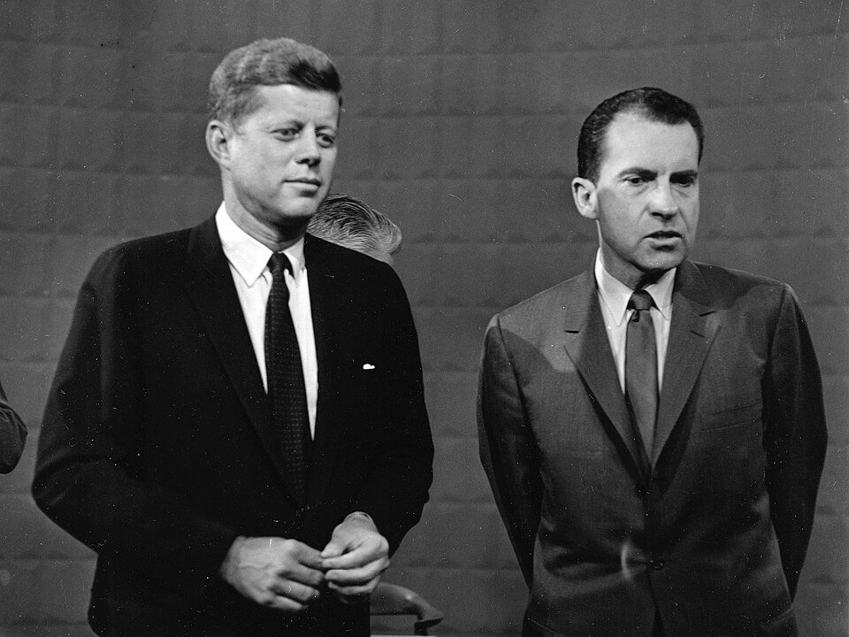 Two men in dark suits standing side by side in a bare television studio, one looking calm and the other drawn.