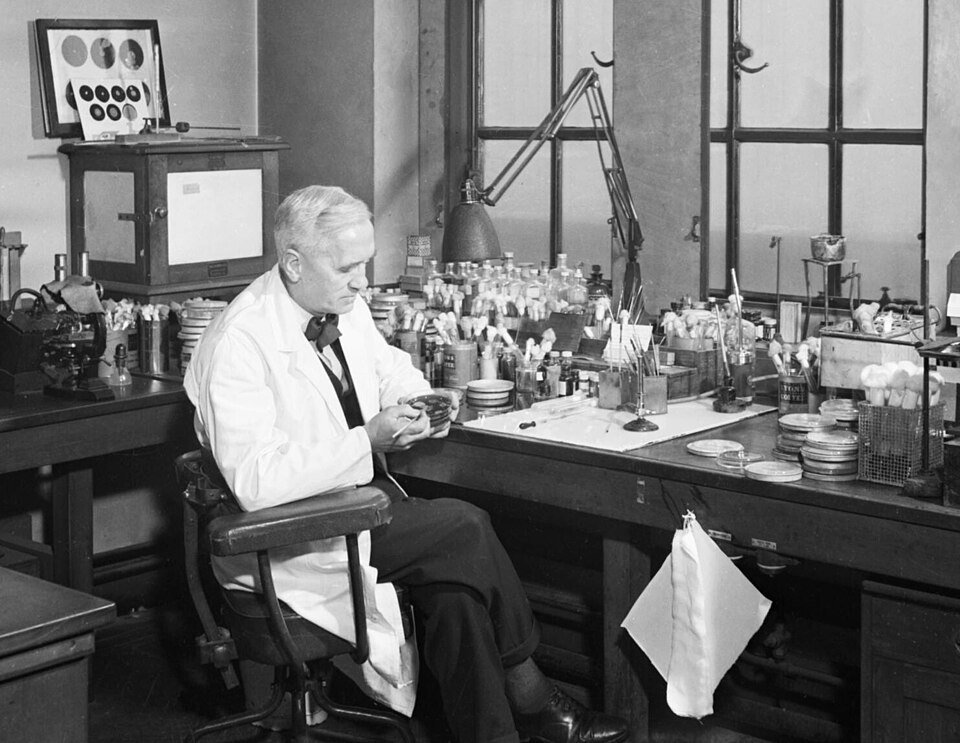 Black and white photograph of an older man in a white coat seated at a cluttered laboratory bench examining a small dish.