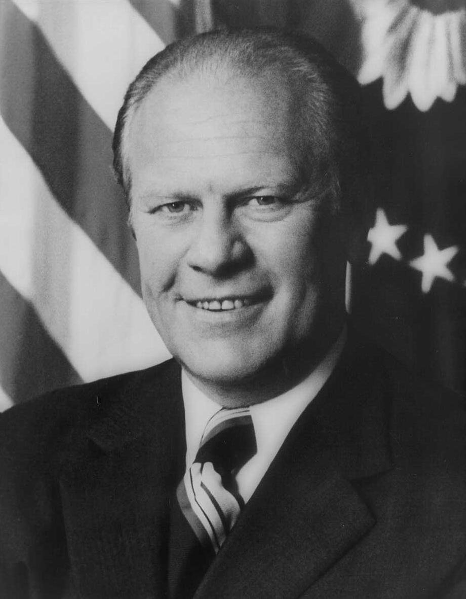 A black and white official portrait of a middle-aged man in a dark suit smiling in front of an American flag.