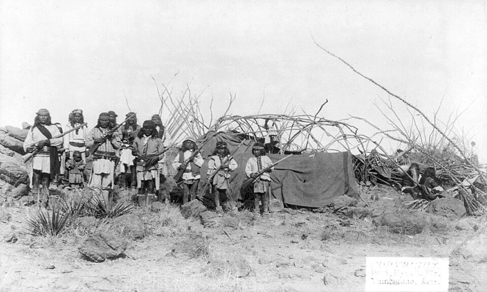 Black and white photograph of a group of Apache men and women seated and standing among scrub, several holding rifles.