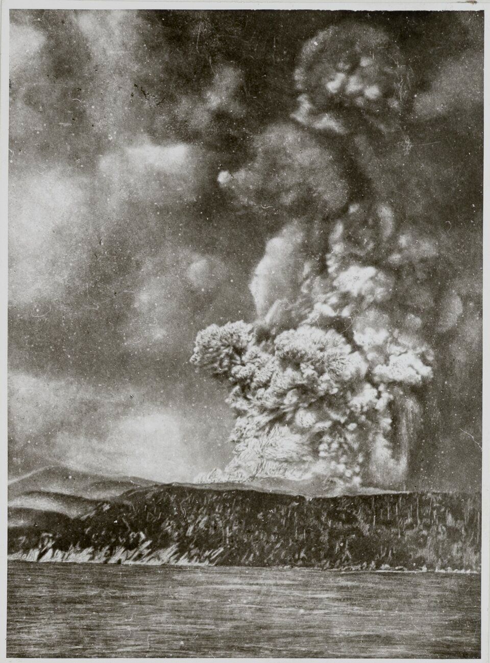 Black-and-white photograph of a huge dark ash cloud erupting from a coastal landform, seen across open water.