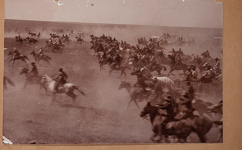 A sepia photograph of hundreds of riders and wagons charging across open prairie in a huge cloud of dust, many blurred by movement.