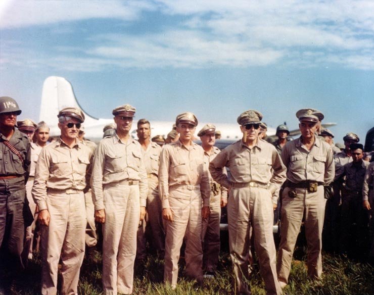 Color photograph of a group of military officers in uniform standing on an airfield beside a large propeller aircraft.