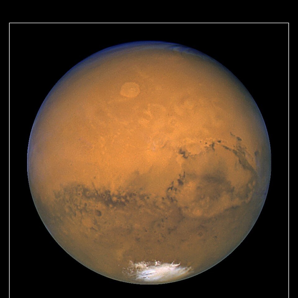 Photograph of the planet Mars, an orange-red sphere with visible surface markings and a polar ice cap, against black space.