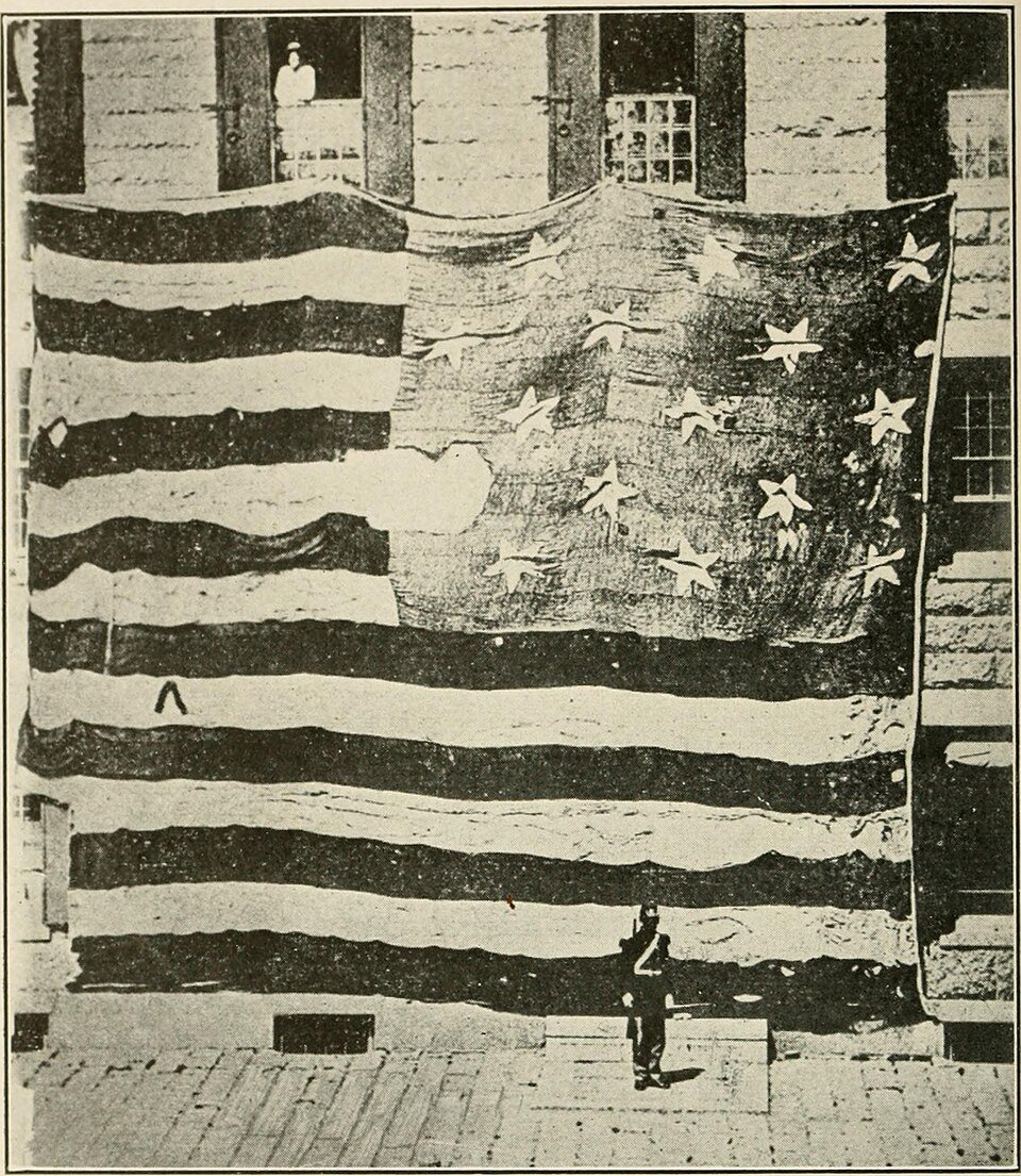 An enormous early American flag with fifteen stars hung across the front of a building, a single sailor standing beneath it for scale.