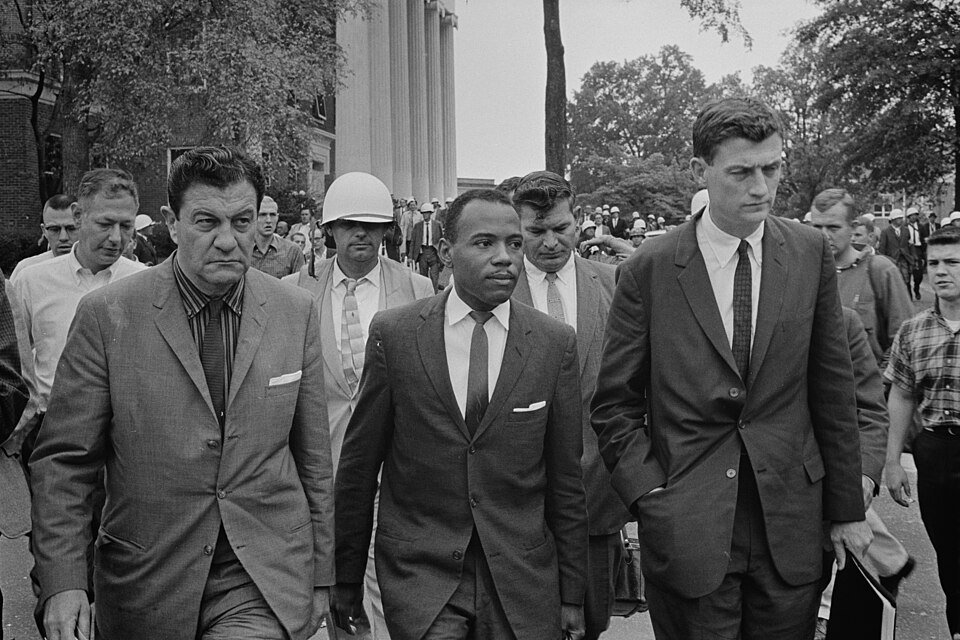 A young man in a suit and tie walking through a crowd flanked by grim-faced men in suits, with helmeted officers behind.
