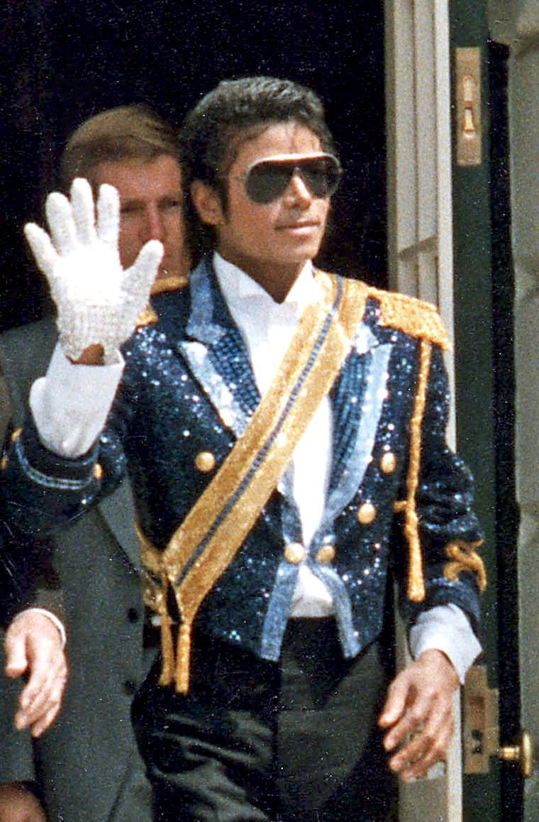 Color photograph of a man in a sequined military-style jacket, sunglasses and one white glove, waving to a crowd.