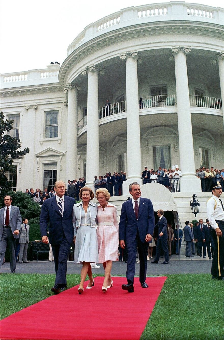 Colour photograph of two couples in formal dress walking a red carpet away from a white columned building.