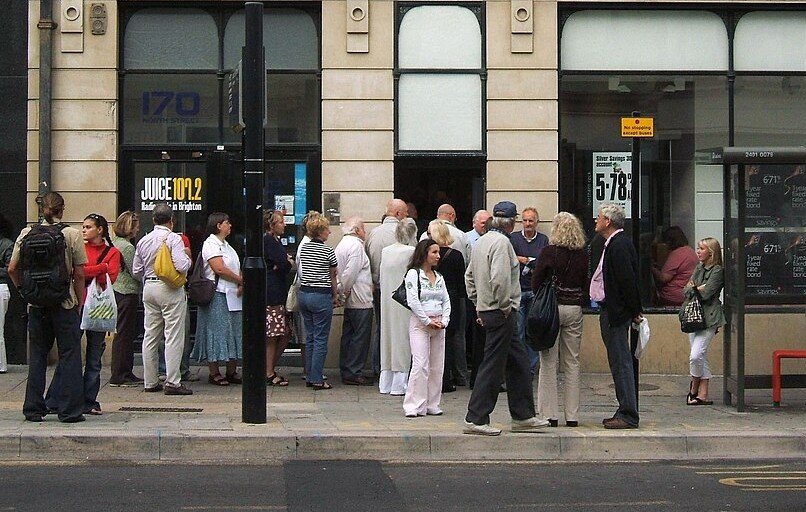 A long queue of ordinary people waiting on a British high street outside a branch with savings rates in the window.