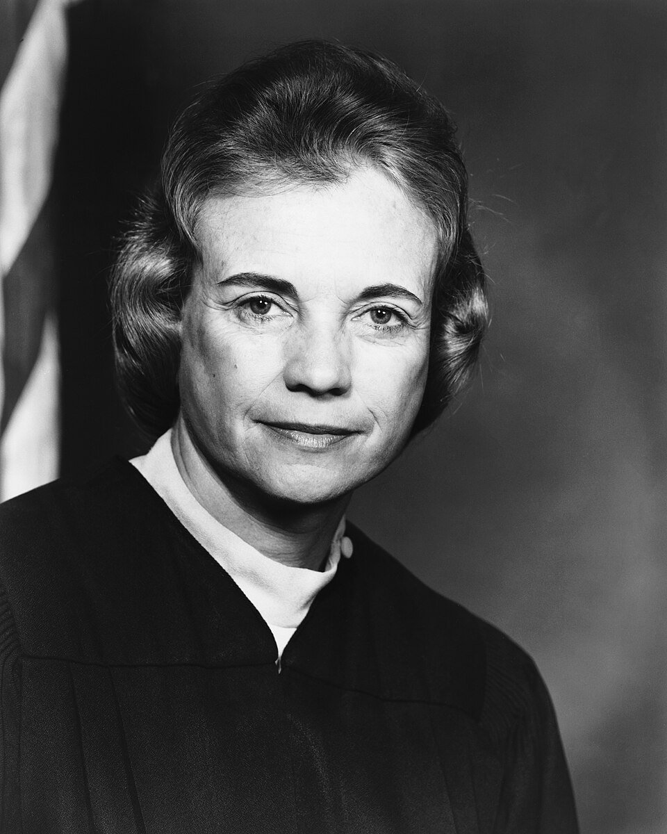 A formal portrait of a woman in black judicial robes in front of a flag.