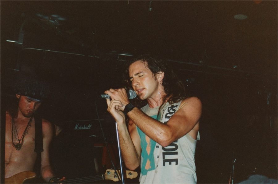 Color photograph of a young rock singer with long hair, eyes closed, gripping a microphone on a dark stage.