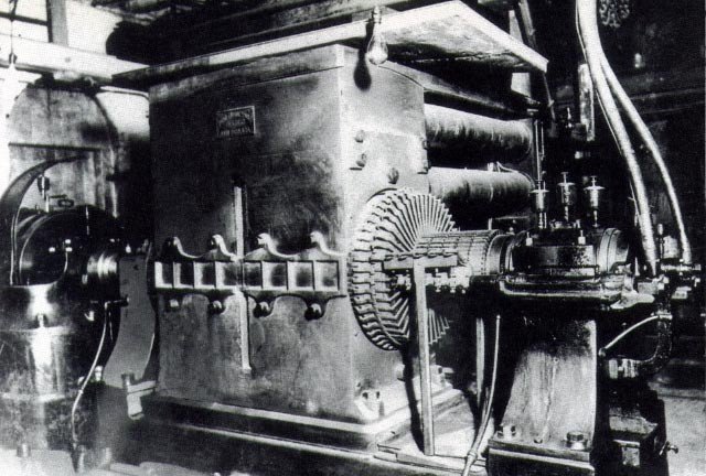 Black and white photograph of a massive early industrial dynamo with heavy pipework in a brick engine room.
