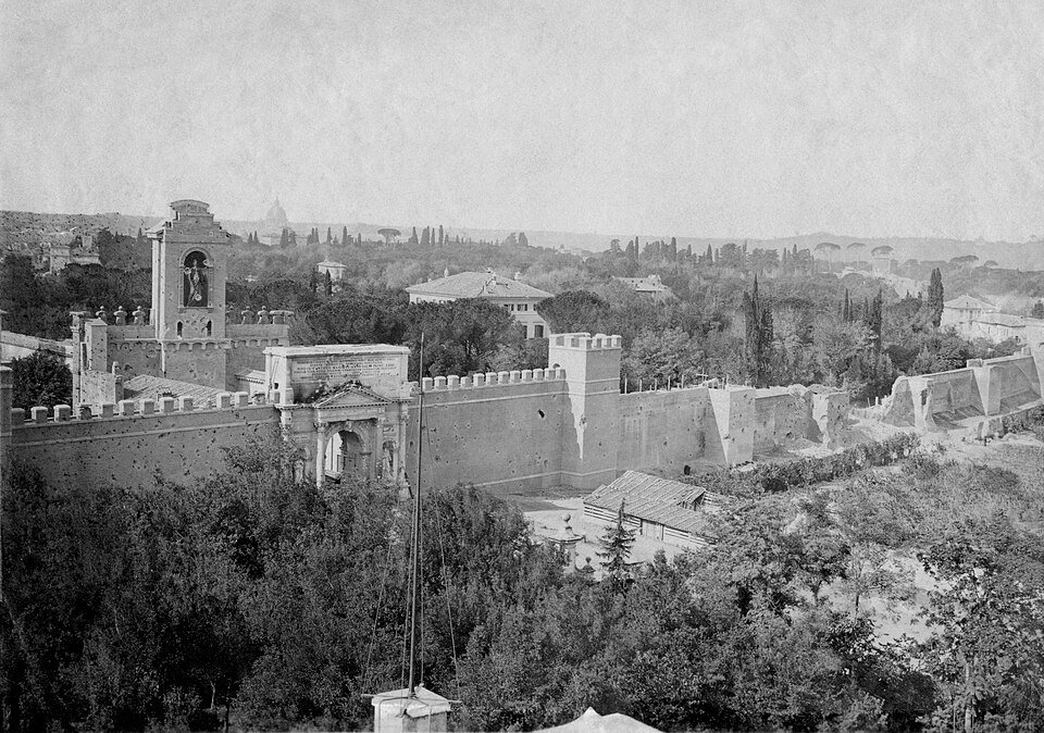 An old photograph of a long city wall pocked with shell damage and broken open near a gate tower, with domes visible in the distance.