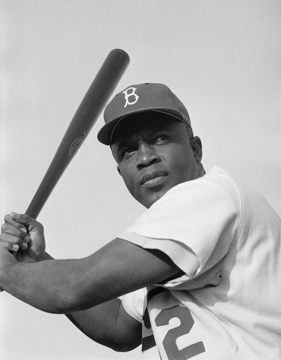 Black-and-white photograph of a baseball player in a Brooklyn cap mid-swing, bat raised over his shoulder.