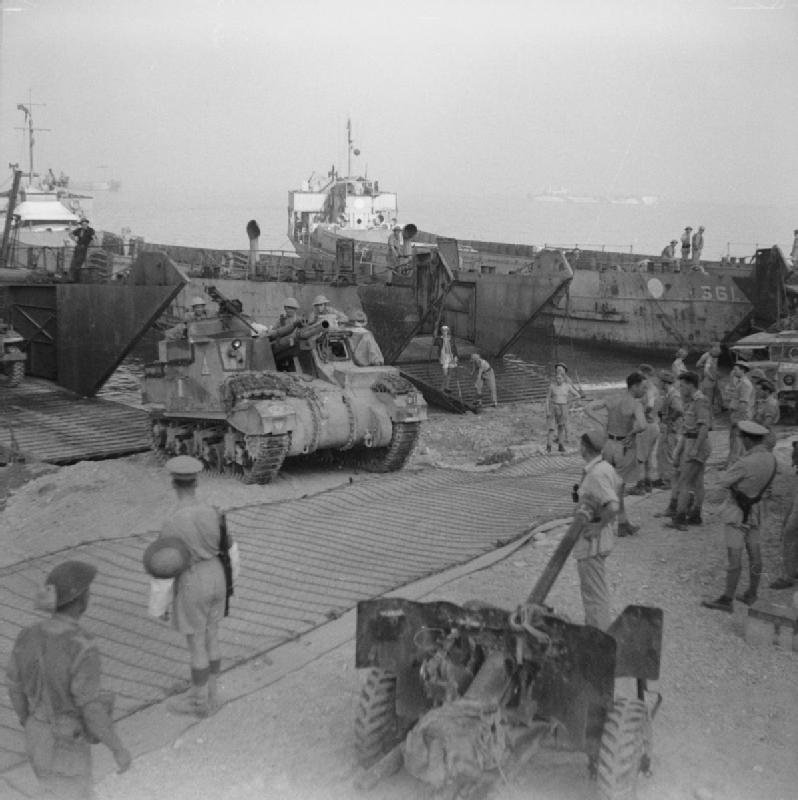 Black and white photograph of tanks rolling off landing craft onto a beach as soldiers watch.