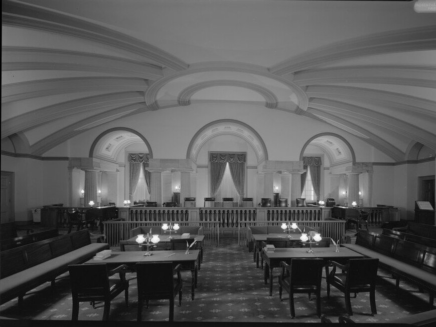 An empty vaulted courtroom with a curved bench, lamps on the tables and rows of benches on either side.