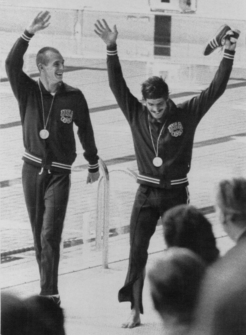 Black and white photograph of two swimmers on an Olympic podium in tracksuits, one raising his arms.