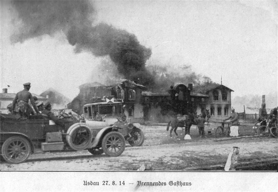 Old photograph of a burning building with heavy smoke, soldiers and horse carts in the foreground.