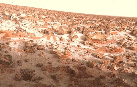 Colour photograph of a rocky reddish plain scattered with boulders and patches of white frost, under a pale sky.