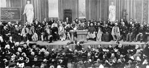 Grainy black and white photograph of a large stage lined with seated dignitaries before a packed hall.