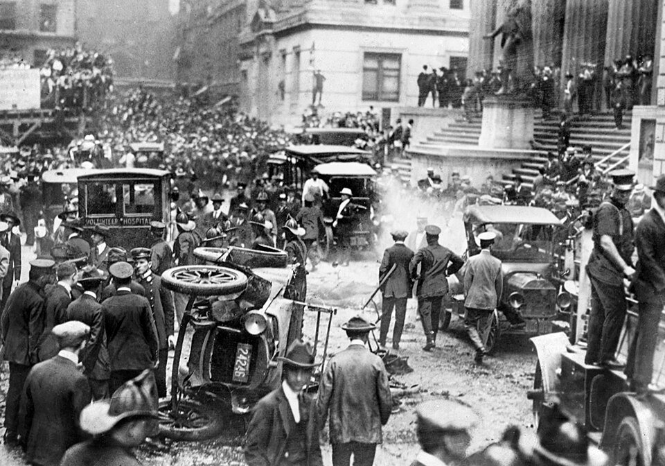 A black and white photograph of a crowded city street with an overturned motor car, smoke, and crowds gathered on the steps of a columned building.