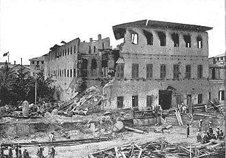 Black-and-white photograph of a badly damaged multi-story building, with rubble in front and exposed columns.
